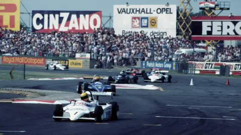 BRDC/Silverstone Archive A white racing car leads the way on a racing circuit with thousands of fans in attendance