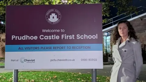 Stacey Martin wearing a grey trench coat and standing by a large purple sign which says "Prudhoe Castle First School". Ms Martin has black hair and is looking at the camera. There is green grass below littered with brown leaves and a school building in the background. It is dark.
