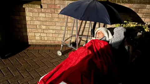 An elderly man lying on a concrete driveway covering in a red blanket and duvets. An umbrella is shielding him from the rain. 