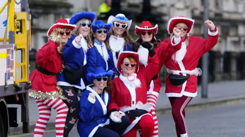 Group image of eight women wearing red and blue santa jackets with cowboy hats and heart-shaped shades raising their hands and cheering for a picture