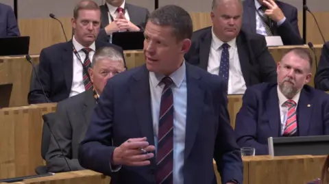 Rhun ap Iorwerth speaking in the Senedd, surrounded by other Members of the Senedd.