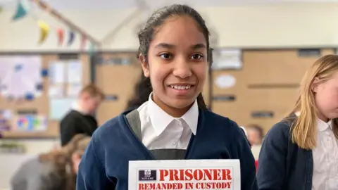 BBC/ Nicola Rees A young schoolgirl with dark hair stands in a classroom holding a card which reads 'Prisoner Remanded in Custody'. She is smiling.