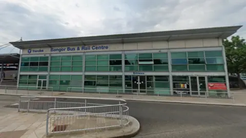 A building with lettering in blue stating: Translink. Bangor Bus & Rail Centre. The word entrance is displayed over a door. The sky is cloudy. 
