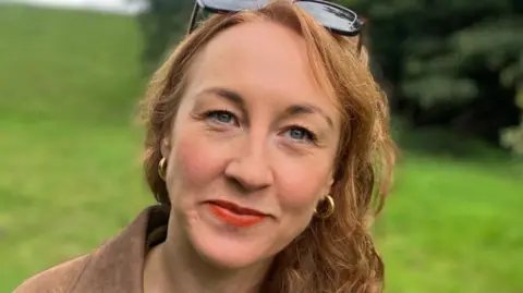 Elizabeth Baines/BBC A woman with long curly red hair smiles at the camera. She is standing in a park
