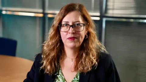 Det Insp Lucy Forde wearing a green patterned blouse under a black jacket, with rectangular framed glasses and gold hoop earrings. She is sitting at a table in an office setting, looking at the camera.
