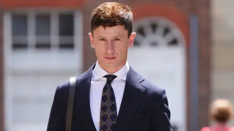 Oisin Murphy, a man with short dark hair and wearing a blue suit, walking along a pavement next to a row of parked cars. He's wearing a suit and looking at the camera.