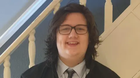 Wearing dark framed glasses, a white shirt, dark tie, and with shoulder length dark brown hair, Ethan Entwistle looks to the camera with an open-mouthed smile. Behind him are the bannisters of a flight of stairs.