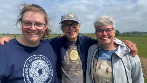 BBC Jane Harrison, Kathryn Murphy and Sarah Miles stand with their arms around each other in a large field. They are all smiling and wearing t-shirts, one with the words Community Archaeology North on it. All have glasses, two have grey hair, the woman on the left is younger and has brown hair tied back. 