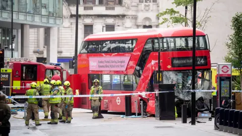 PA Media A red double-decker bus with its front crushed and windows broken. Firefighters in yellow helmets are surrounding the scene, and a fire engine is parked nearby.