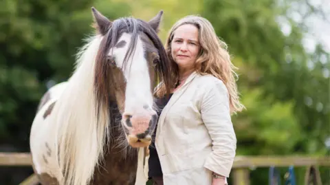 The lady stands close to the horse, as they both look into the camera. The lady has long wavy hair and is dressed in neutral colours. The horse is a similar height to the lady and is brown and white in colour. They are standing in a paddock and there are lots of green trees surrounding them.