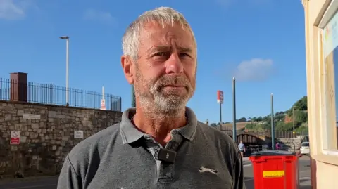 A man wearing a grey shirt standing outside of a train stop. Blue skies behind.