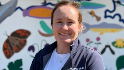 Hannah Gutteridge, Thames Path manager, wearing a fleece which says "Thames Path" on it and in the background is the mural with all of the images of wildlife painted on it.