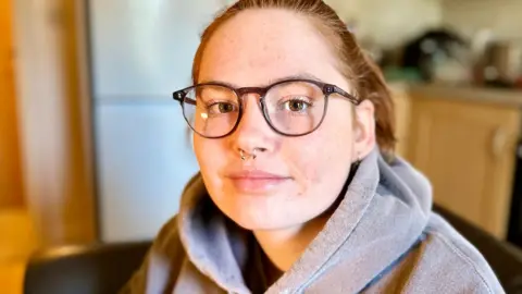 Head and shoulders portrait photograph of Stevie, taken in her kitchen, although the background is blurred. Stevie has auburn hair, tied back into a ponytail and brown glasses on. She has a nose piercing and is wearing a grey hoodie.