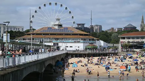 Bournemouth Pier on a sunny day. You can see a big wheel behind a building with solar panels on the roof. There are a lot of people on the beach and on the pier. There is sand and you can see the sea.