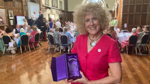 BBC Ms Barlow wears a bright fusia dress and has blond curly hair in a bob. She holds the glass award which sits in a purple satin box. The volunteers sit around tables in the background within the wood panelled room of the Guildhall.