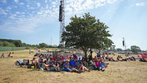 PA Glastonbury Festival attendees sitting under a tree in the shade on a sunny day