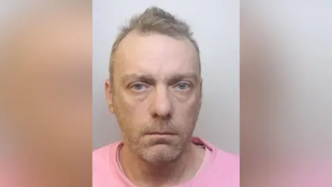 A police mugshot of Dean. He has stubble, light brown short hair and is wearing a pink T-shirt.