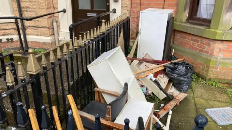 A pile of waste - mostly broken up wooden furniture, is dumped in the forecourt of a terraced house, beside the fence it shares with another house. There are bits of wood, a chair, and a black bin bag.