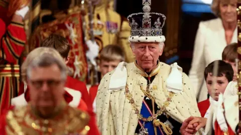 King Charles III is wearing the Imperial State Crown and the Robe of State as he leads the royal procession in the House of Lords. His left hand is held and he has four page boys dressed in red and white to his side.
