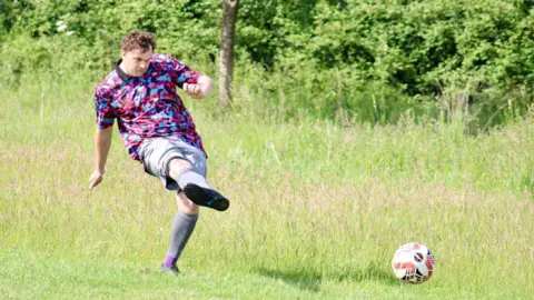 A man in brightly coloured sports shirt kicking a football while playing on a grass field, with trees and greenery in the background. 