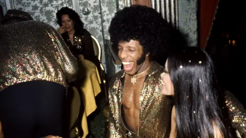 Getty Images Sly Stone and his newlywed wife Kathy Silva grin and laugh, dressed in gold. She has confetti scattered through her hair.