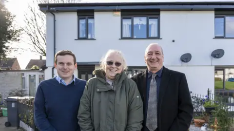 City of Wolverhampton Council A man on the left is in a blue jumper, the woman in the middle is wearing a green coat and the man on the right is wearing a dark jacket, a tie and a shirt. The house behind them is white and they are looking at the camera. 