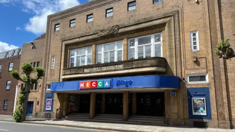 Historic brick building with extensive brick work. It has giant windows and some smaller ones above a big blue sign. It says "MECCA Bingo". The building is on a main road with a tree outside the big stepped entrance.