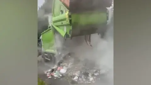 A green bin lorry is partially opened up as waste is deposited on the road side with smoke pouring from the vehicle. 