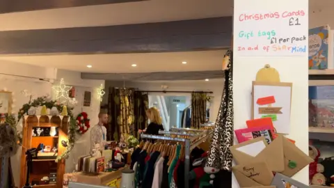 View inside Space4Autism's shop. There is a sign on a pillar in the foreground that says "Christmas Cards £1" and the top and in the background staff are stood behind the till and rows of clothes on hangers.