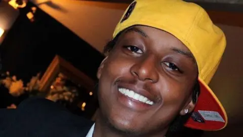 PA Eddie Kinuthia, a young man, is smiling. He is wearing a yellow cap backwards and a diamond ear stud.