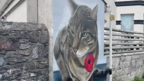 BBC A painting of a tabby cat, with a red poppy and a blue background, on a wall of a house.