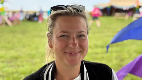 A woman with blonde hair tied back and sunglasses on her head. She is sitting down with grass in the background and a blue and purple flag billowing behind her.