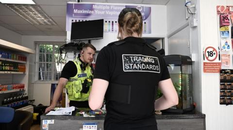 A male police officer standing at a vape shop counter with the back of a female Trading Standards officer in front of the desk