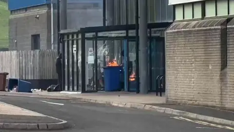 A blue wheelie bin being brought into a building, it is on fire.