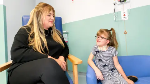 Ashley and Macie are sat next to one another on hospital chairs. They are looking at each other and smiling.