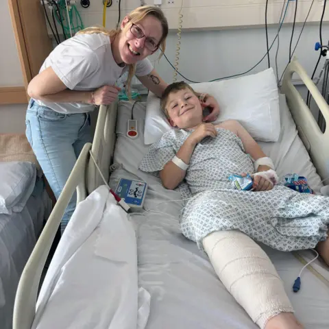 Jo Newman Jo Newman, who has blonde hair and glasses, with her son at Cardiff's Noah's Ark Children's Hospital. He is lying in a bed with a bandaged leg in a hospital gown