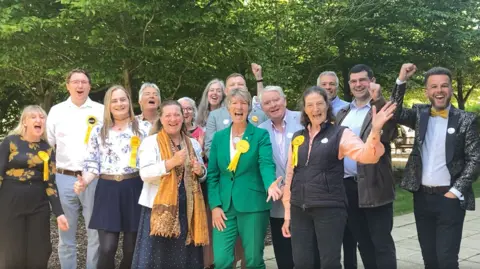 Emma Howgego/BBC A group of Liberal Democrats standing outside and cheering. They are wearing yellow ribbons and have their hands out and up in the air. THey are all smiling.