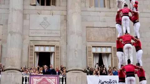 La Mercè Festival Acrobats in red tops and white pants stand in a human pyramid while people stand on a nearby balcony watching and clapping. 