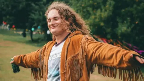 Danny smiles at the camera as he holds his arms in a cross position. He has a drink in a can in one of his hands and he's wearing a light brown jacket with tassels and a grey t-shirt. Danny has shoulder-length curly hair which is light brown. 