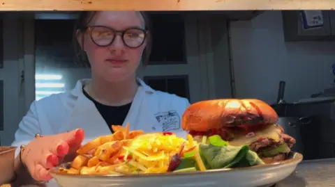 BBC/Seb Cheer A young woman, Lucy, places chips onto a plate of food, under a hatch in a commercial kitchen.