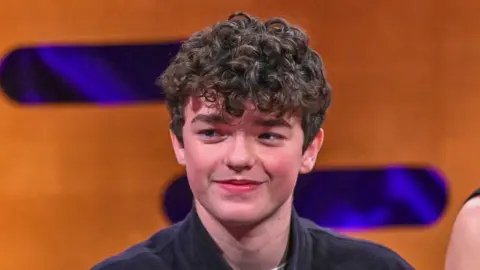 PA Media Owen Cooper smiles during Graham Norton interview. He has a mop of dark curly hair and wears a dark shirt. The backdrop is an orange wall decorated with purple lozenge shapes. It is a head shot of him. 