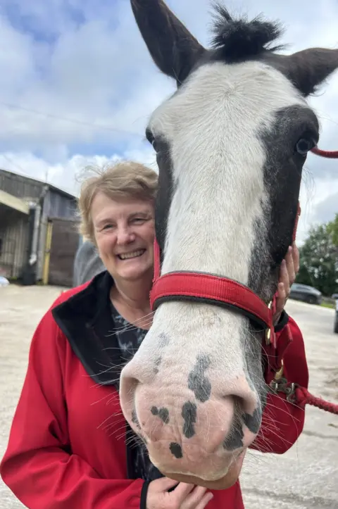 Margaret Haes Riding Centre Debra Batchelor smiles at the camera as she stands with a black-and-white horse leaning over her shoulder. Debra has short blonde hair and is wearing a red jacket.