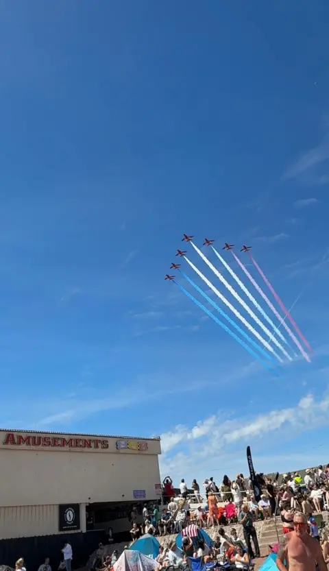 Seven Red Arrows planes fly in formation over Paignton seafront in a blue sky with a few white fluffy clouds. There is a crowd of people gathered on the beach looking up at the sky