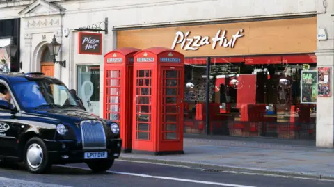 Getty Images A black cab taxi drives past a Pizza Hut restaurant, which has booth tables set against large floor-to-ceiling windows that look out on to the street