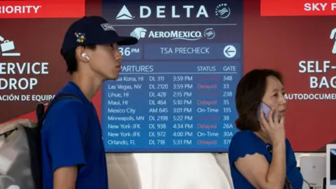 People stand in front of airport sign showing delayed Delta flights 