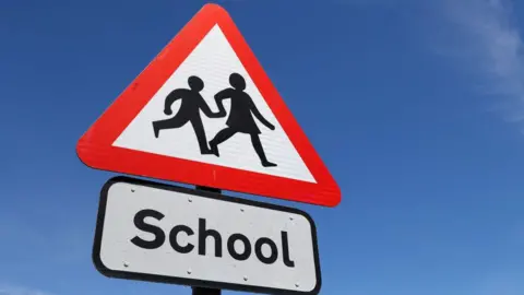 A road sign set against a blue sky. The sign shows the shadow of two children holding hands and walking on a white background, surrounded by a red triangle. The sign underneath reads 'school' in black font on a white background.