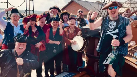 Auckland Shanty Singers The Auckland Shanty Singers, with leader Christopher Miller (bottom left), cheering and dressed as pirates. There is a group of 11 people, plus one dog, on an old style ship.