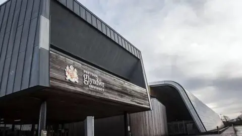 Wrexham University The building prominently displays the text “PRIFYSGOL glyndŵr UNIVERSITY” along with a colorful university crest on a weathered wooden facade.
The structure has a contemporary design, featuring dark metal cladding with sharp geometric lines.