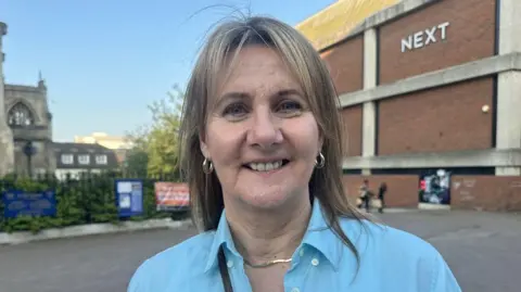 Zoe Dennis/BBC A woman looking towards the camera, smiling. She has dark blonde shoulder-length hair and is wearing a light blue shirt and a gold necklace. She is standing in front of a wall that has the "Next" sign on it.