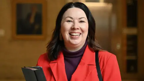 Getty Images Monica Lennon wearing a bright red blazer over a dark turtleneck top stands indoors holding a black folder. The background is softly blurred, with framed artwork and warm lighting visible.